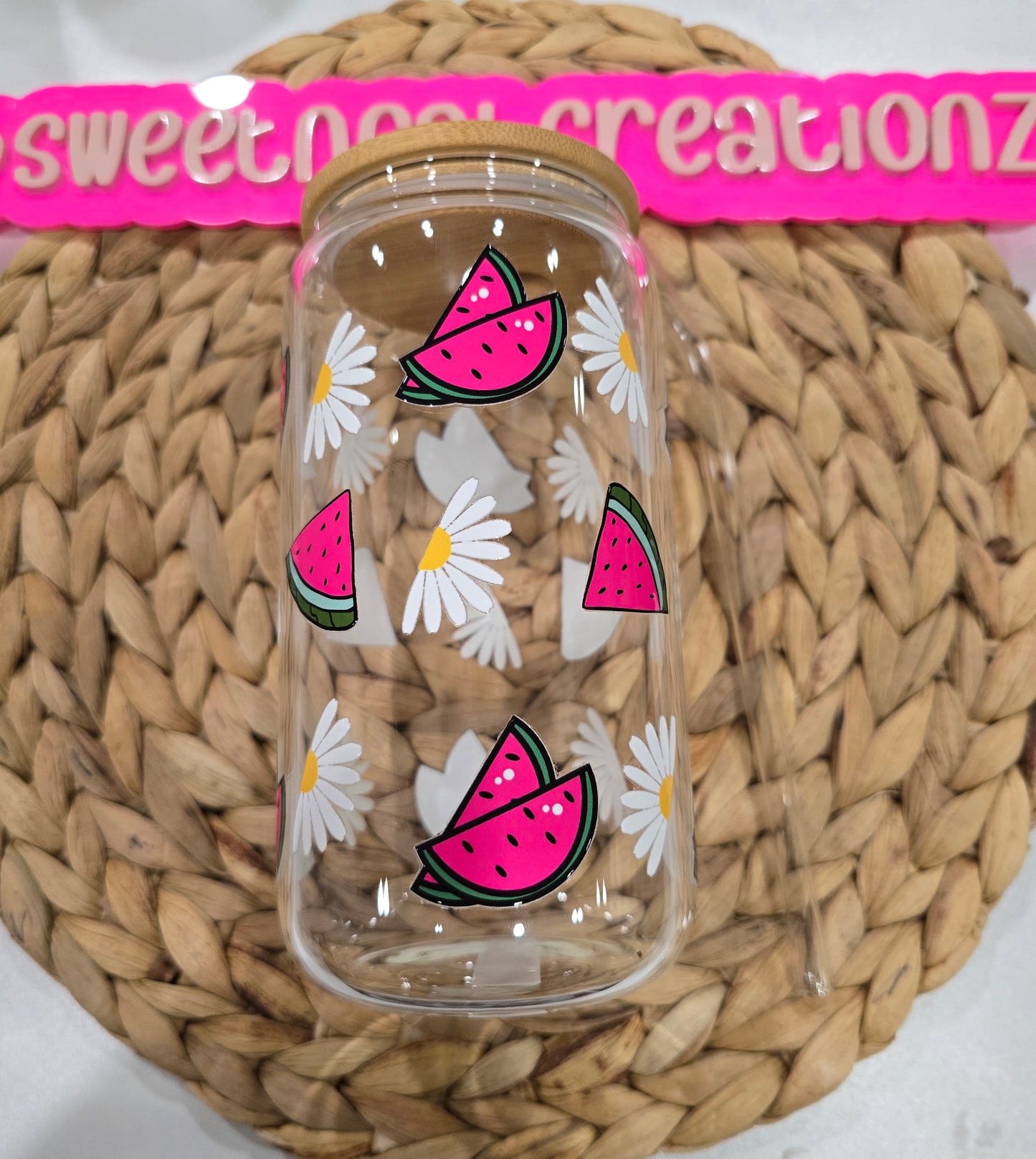 Clear glass jar with pink watermelon and white daisy designs on a woven surface branding.