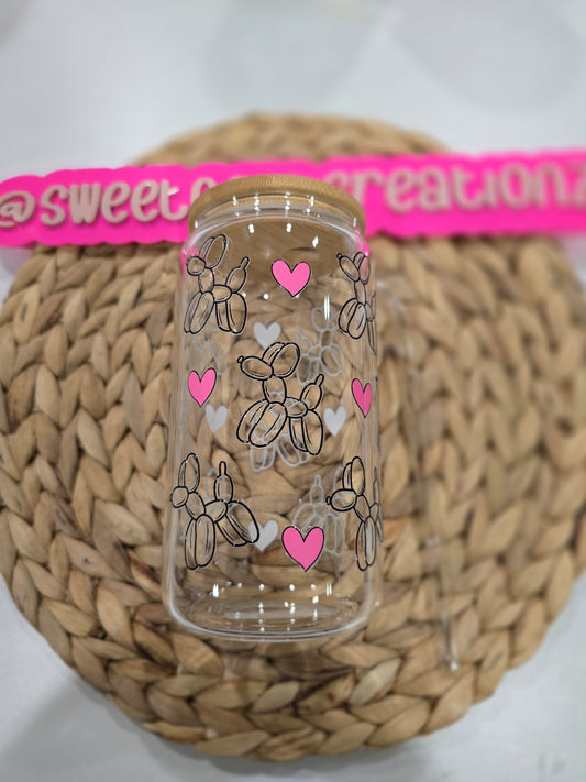 Clear glass tumbler with balloon dog and heart designs on a woven surface.
