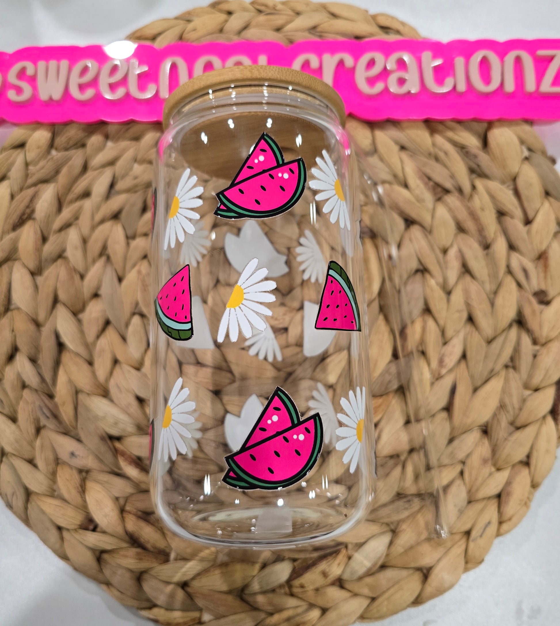 Clear glass jar with pink watermelon and white daisy designs on a woven surface branding.
